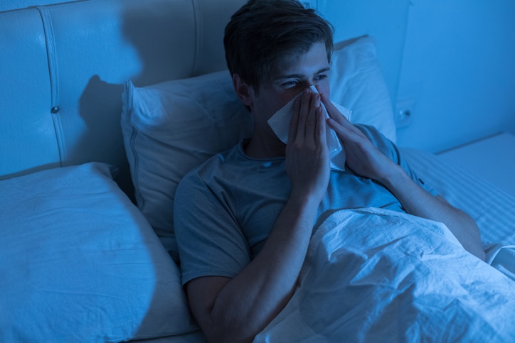 Young man lying in bed and blowing his nose.